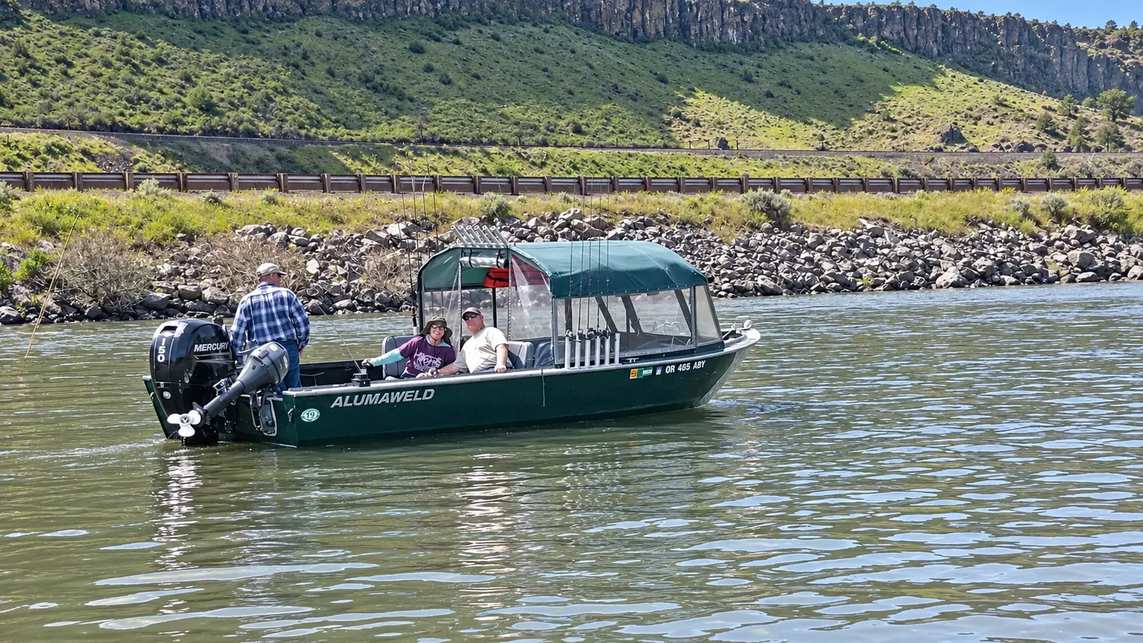 Green Alumaweld Columbia with its cabin canopy up beside a rocky shore, guests on board