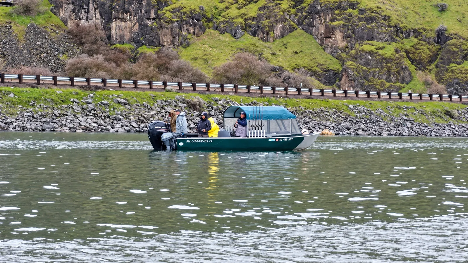 Green Alumaweld Columbia cabin boat on the river with guests aboard and a rail line along the far shore
