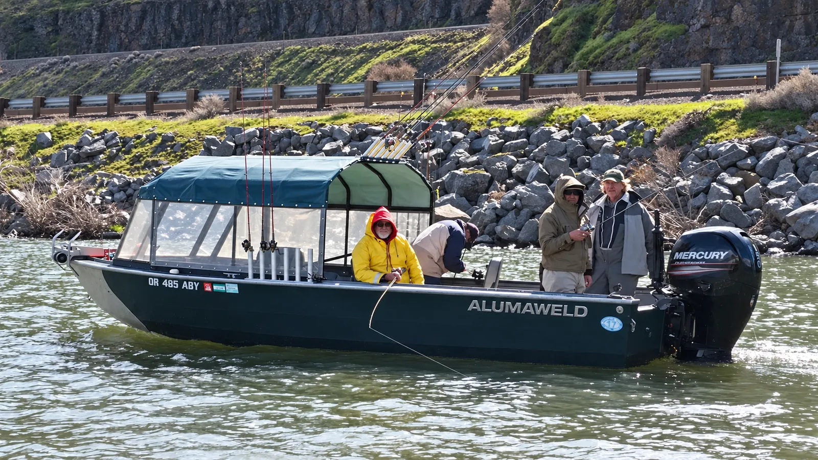 Dark green Alumaweld cabin boat with a Mercury outboard and anglers fishing along a rocky shore