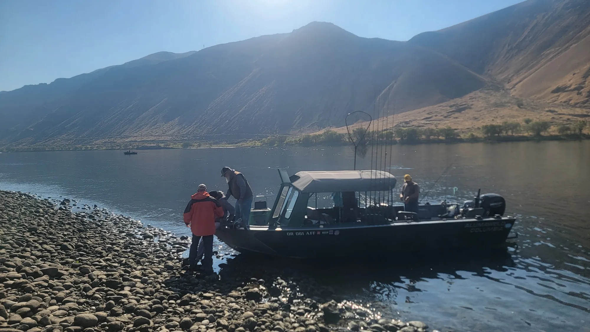 Guide boat with a canopy at a gravel shore below sunlit hills, guests stepping aboard