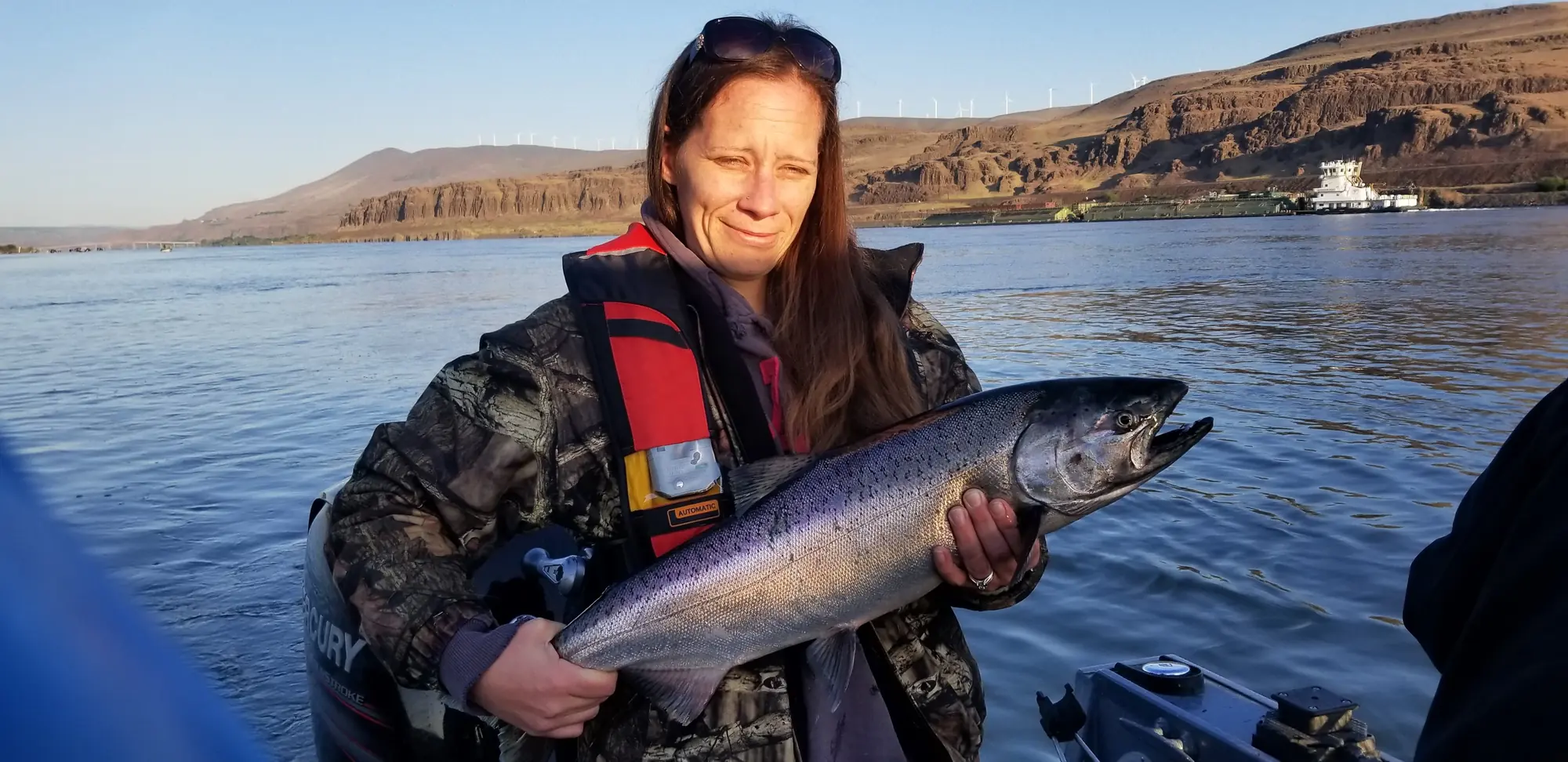 Woman in a life jacket holding a bright Chinook salmon on the Columbia River at sunrise, with a tug and barge behind her
