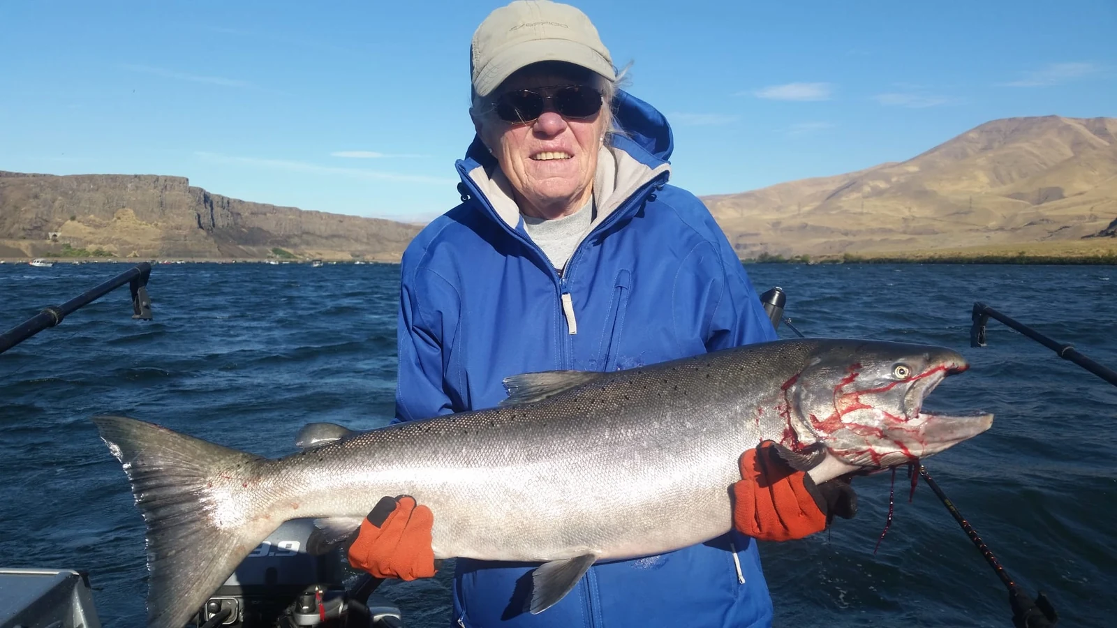 Man in a blue jacket and orange gloves holding a Chinook with brown hills behind him