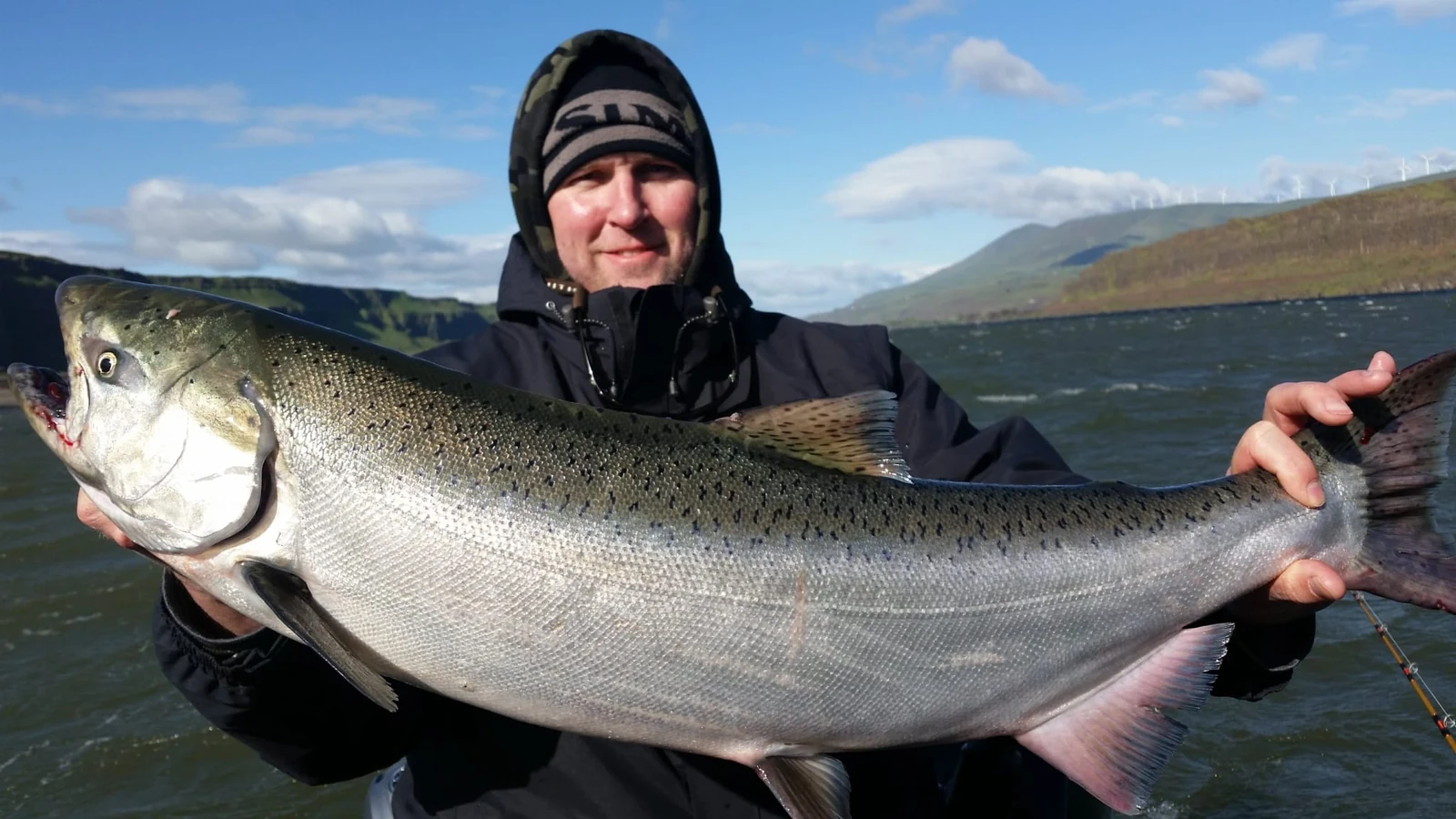 Angler in a hood holding a bright spring Chinook, dry hills behind him