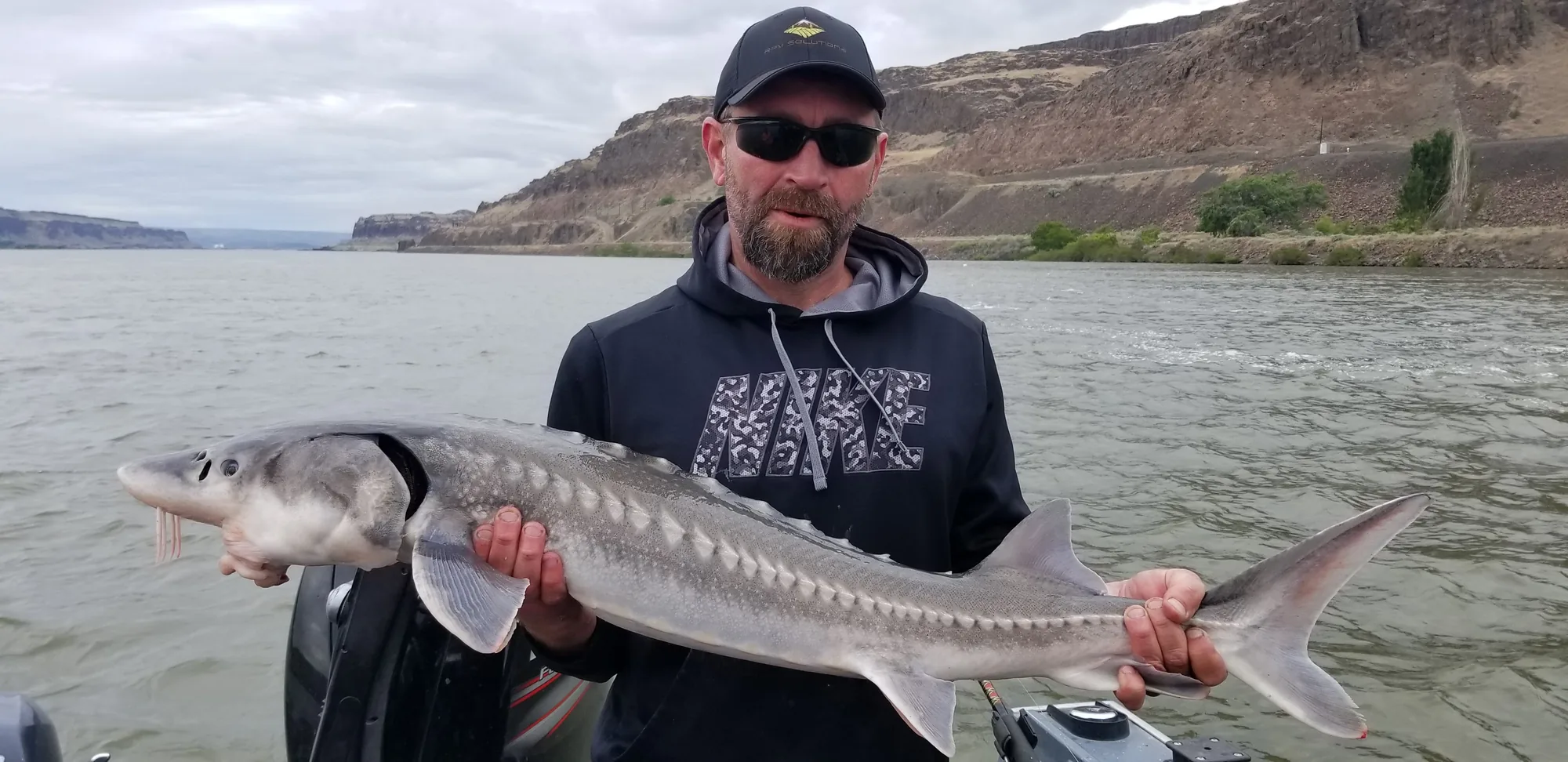 Angler in a black hoodie holding a keeper sturgeon with basalt hills behind him