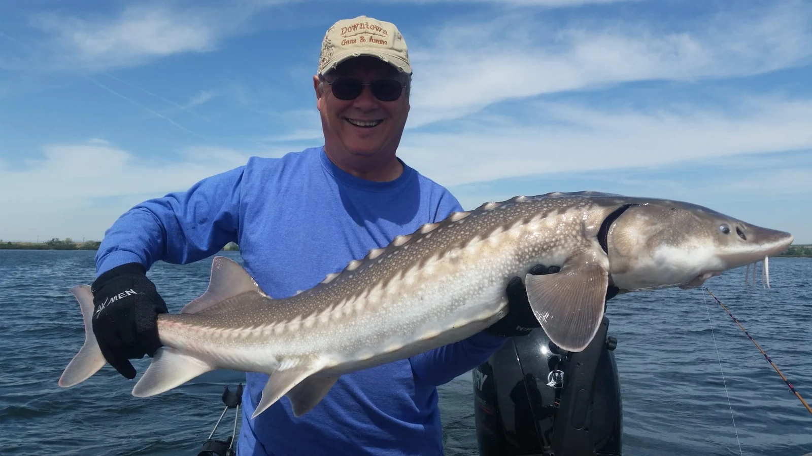 Smiling man in a blue shirt and tan cap holding a keeper sturgeon