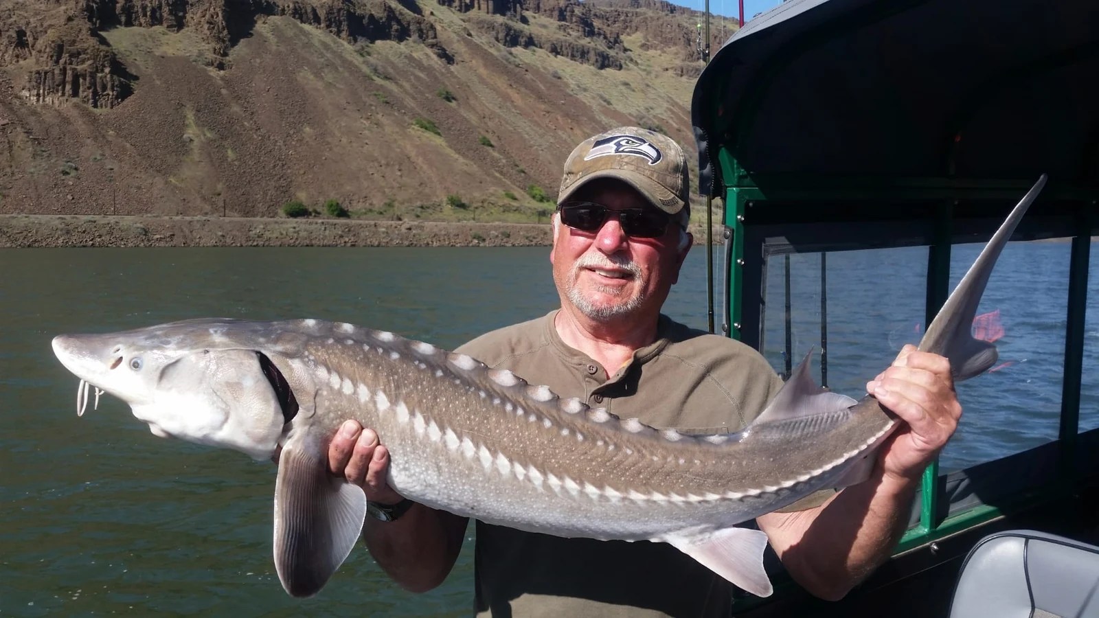 Man holding a keeper sturgeon in the cabin boat, basalt cliffs behind him
