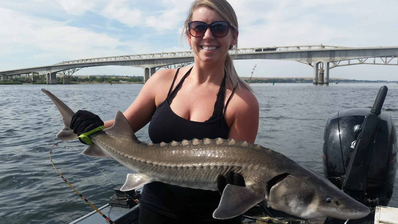 Woman in sunglasses holding a keeper sturgeon with a long bridge behind her