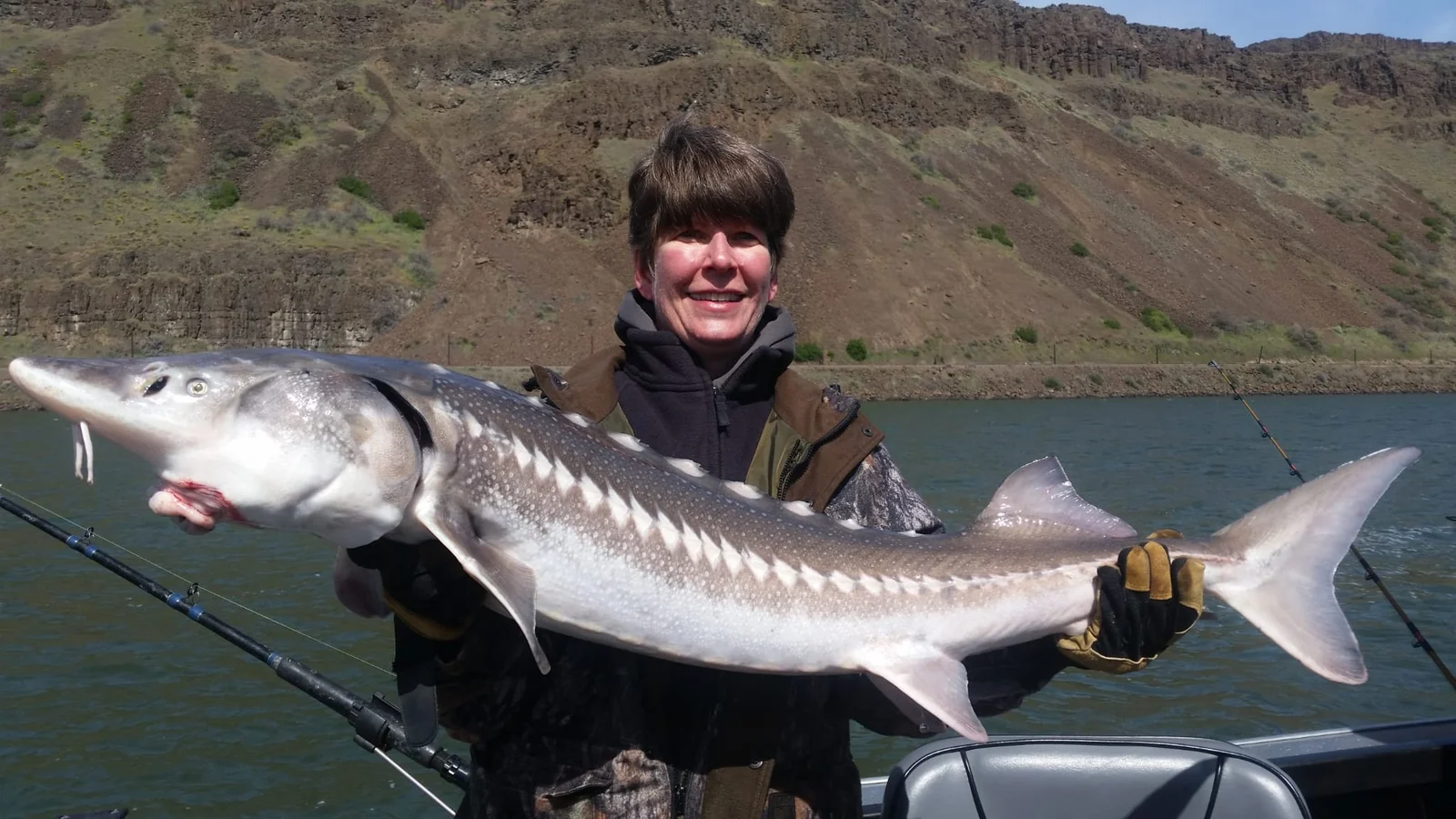 Woman in a brown jacket holding a keeper sturgeon, basalt cliffs behind her