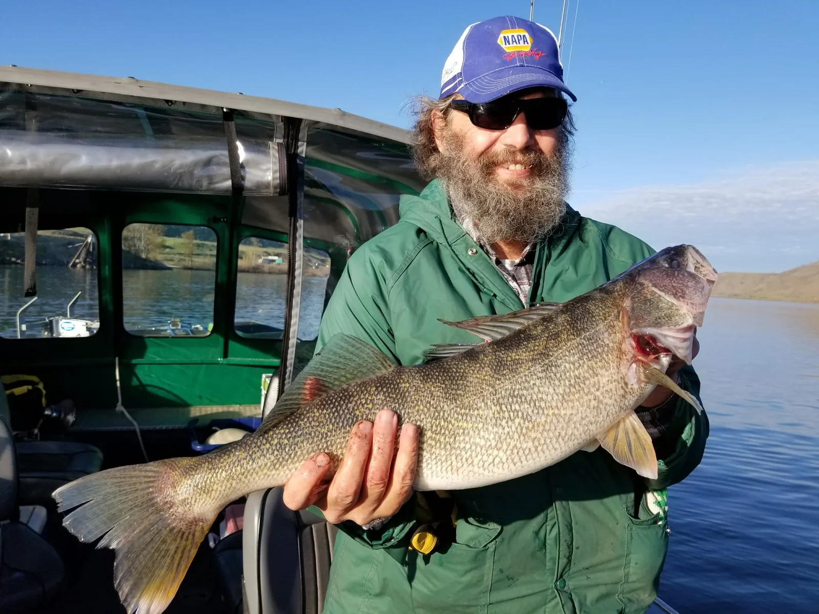 Bearded angler in a green jacket holding a walleye inside the cabin boat