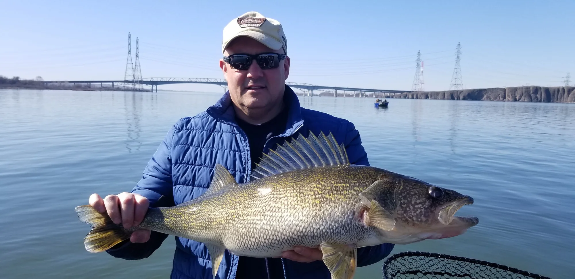 Angler in a blue jacket holding a walleye in both hands, with a bridge behind him