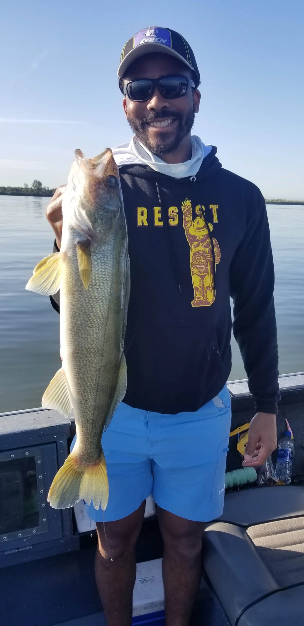 Smiling angler in a black hoodie and sunglasses holding up a walleye